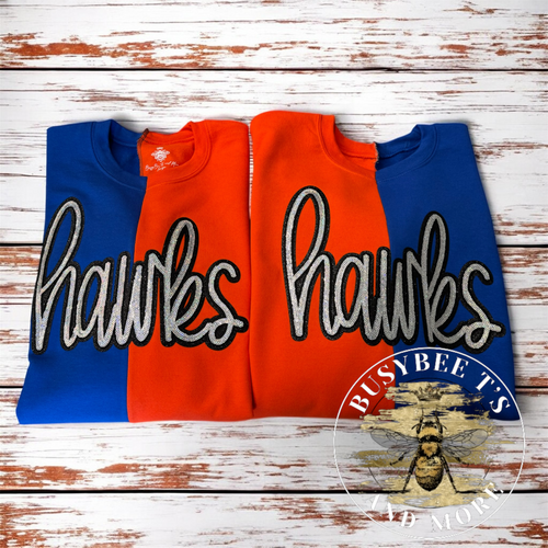 Two folded sweatshirts with a blue and orange vertical split design, each featuring the word 'hawks' in large white script outlined in black across the front, displayed on a distressed white wood background. A circular 'BUSYBEE'S FAD ATO' logo is visible in the bottom right corner.