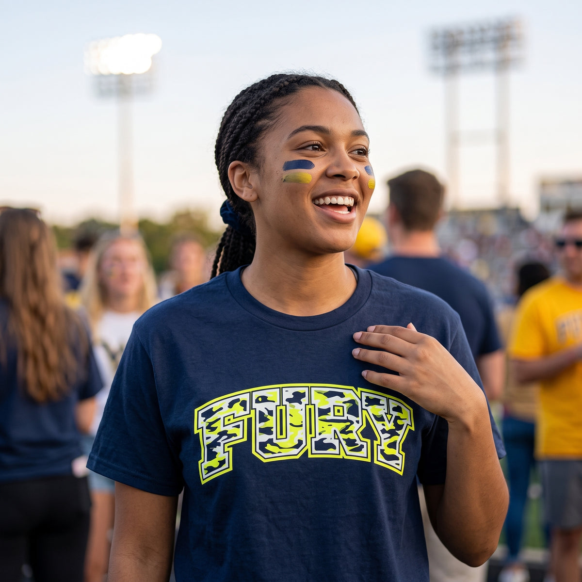 A smiling woman at an outdoor event wearing a navy blue t-shirt with 'FURY' printed across the chest in a yellow and camouflage pattern, with blue and yellow face paint on her cheeks.