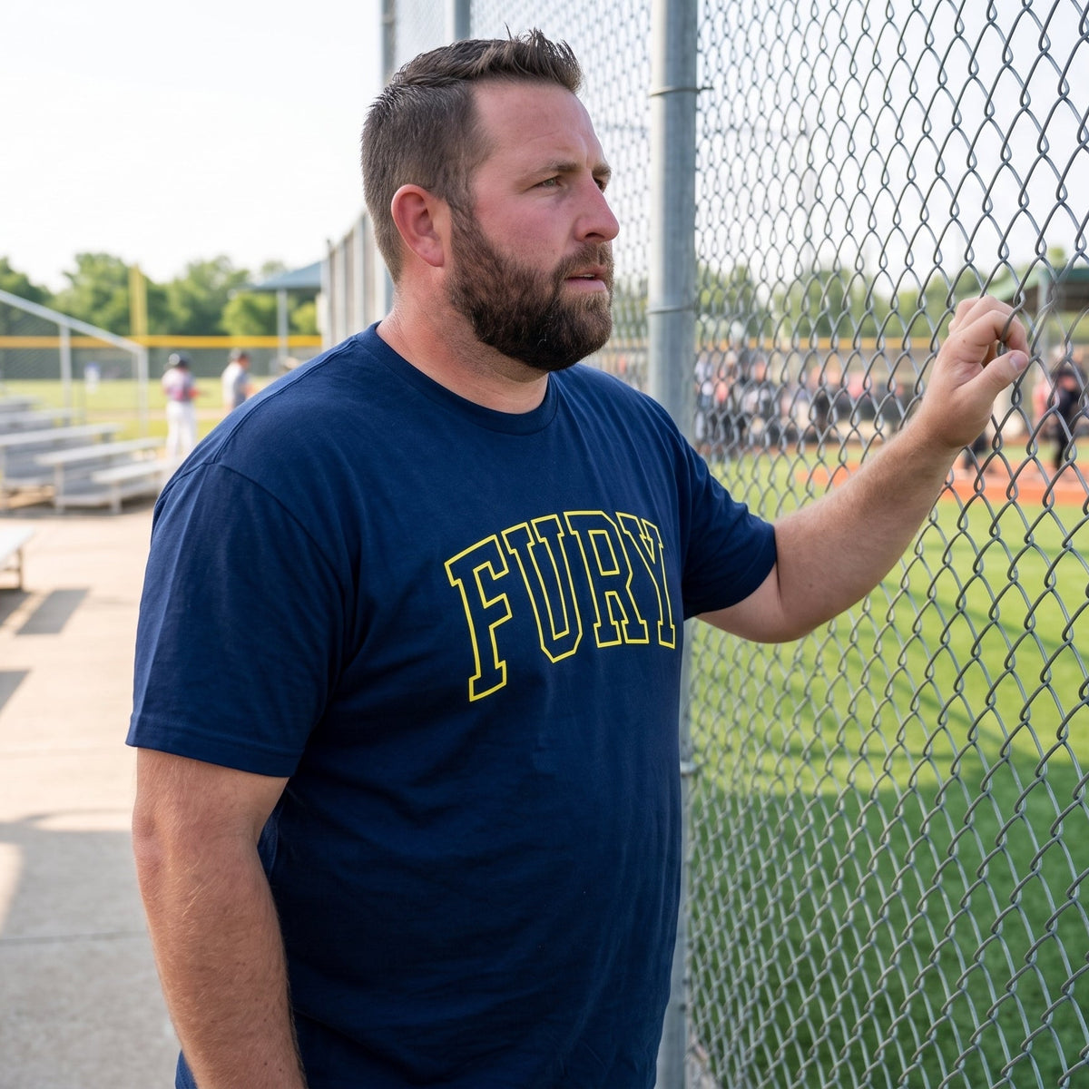 A bearded man in a navy blue t-shirt with 'FURY' printed in yellow across the chest, standing behind a chain-link fence at a baseball field.