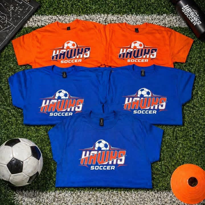 Four custom 'Hawks Soccer' t-shirts in orange and blue are arranged on a green turf field background with soccer-themed props including a ball, whistle, cone, and clipboard.