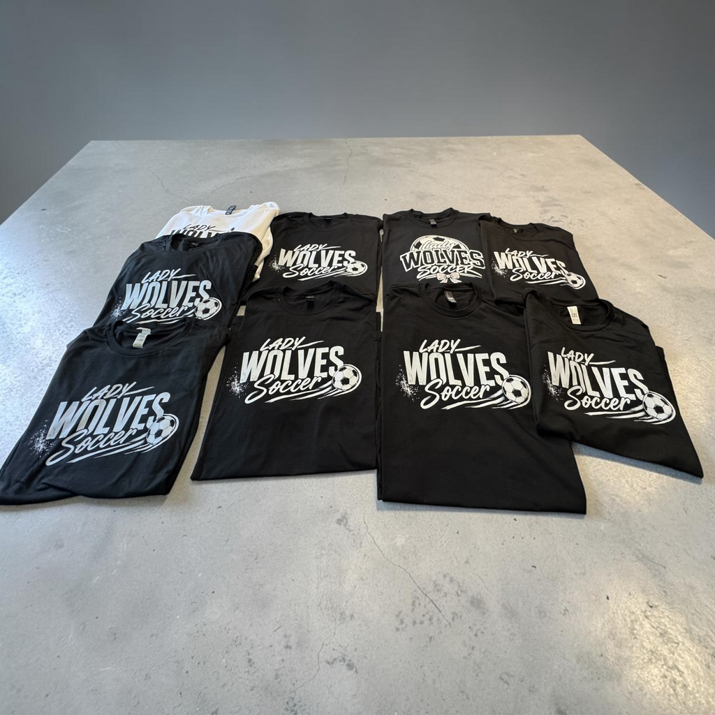 Six black t-shirts and one white t-shirt, all featuring 'Lady Wolves Soccer' printed in white across the front, neatly arranged on a gray concrete surface.