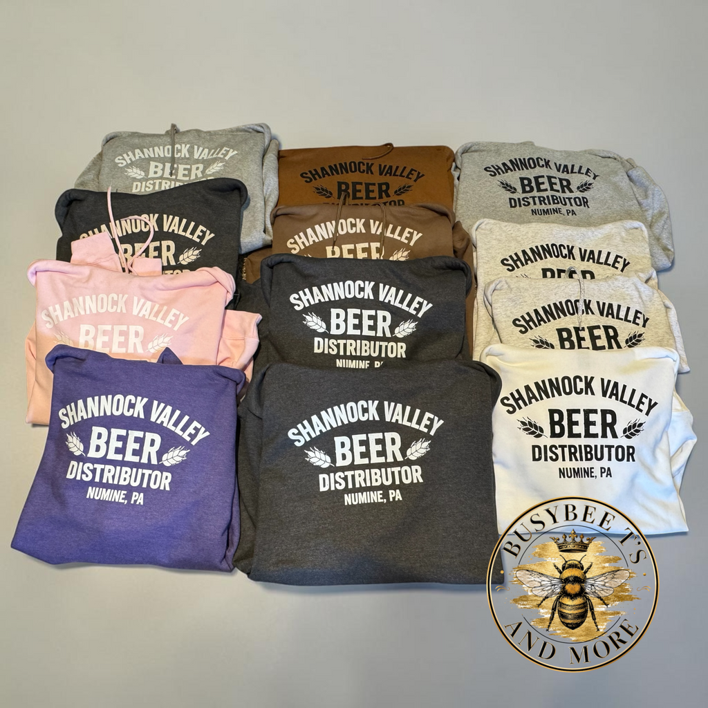 Twelve folded T-shirts in assorted colors arranged in a grid on a light gray background. Each shirt features a white printed graphic that reads 'Shannon Valley Beer Distributor' with a small map and wheat ears illustration.