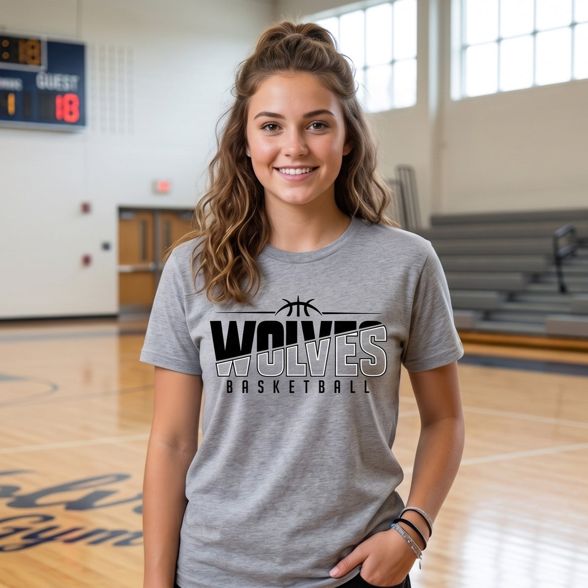 A smiling young woman stands on a basketball court wearing a heather gray t-shirt with a black and white 'WOLVES BASKETBALL' graphic on the front.