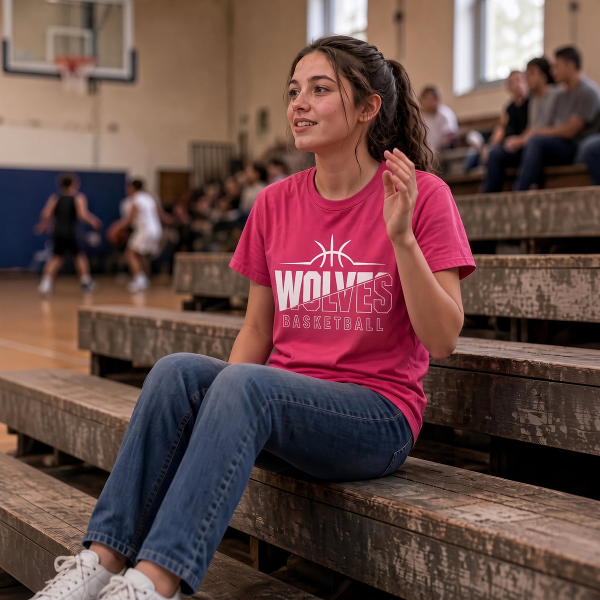 A woman sits on wooden gym bleachers wearing a bright pink t-shirt with a white 'WOLVES BASKETBALL' graphic, in a basketball court with players blurred in the background.