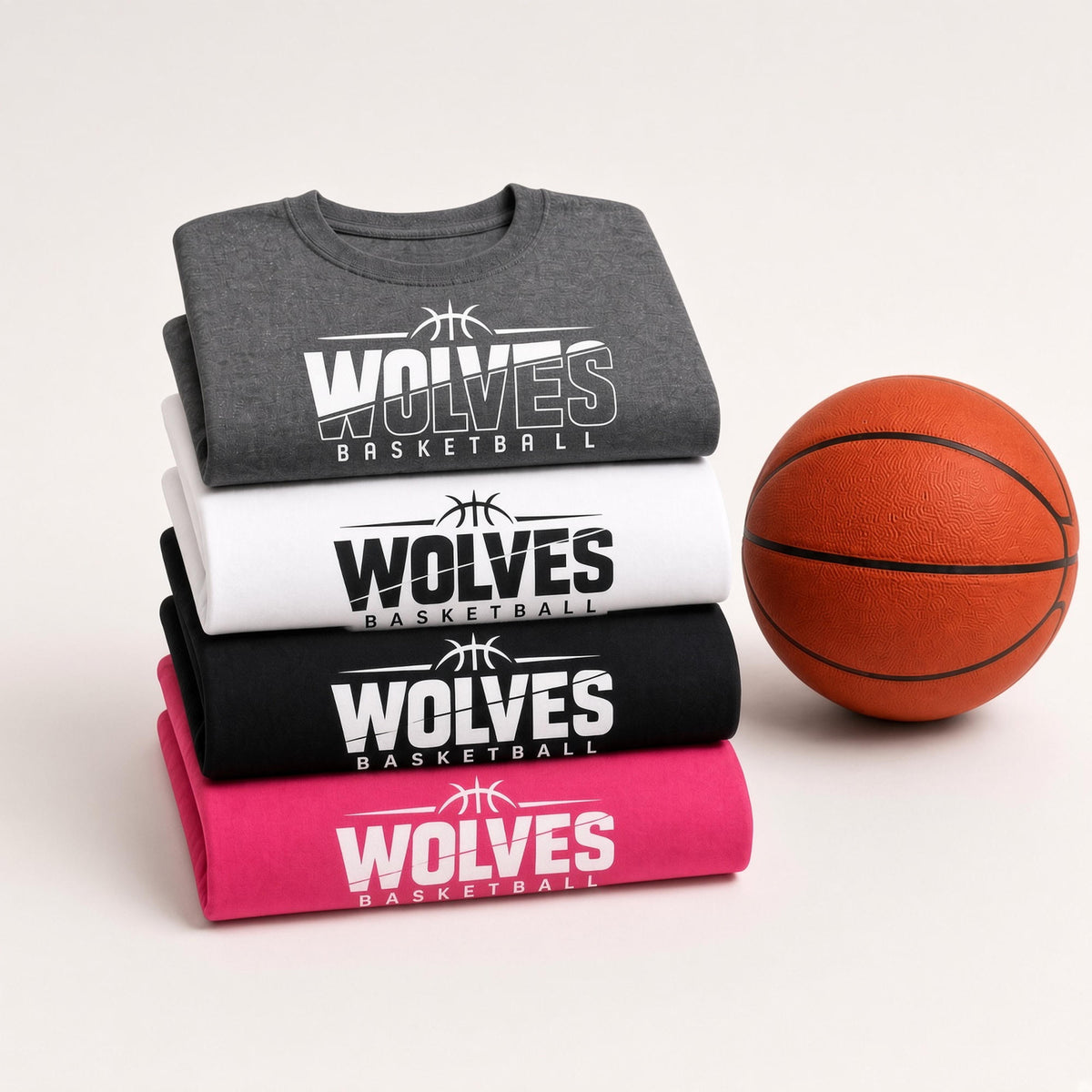 Four folded t-shirts in gray, white, black, and pink are stacked neatly on a light beige background, next to a basketball. Each shirt features a bold graphic print that reads 'WOLVES BASKETBALL' with a basketball logo.