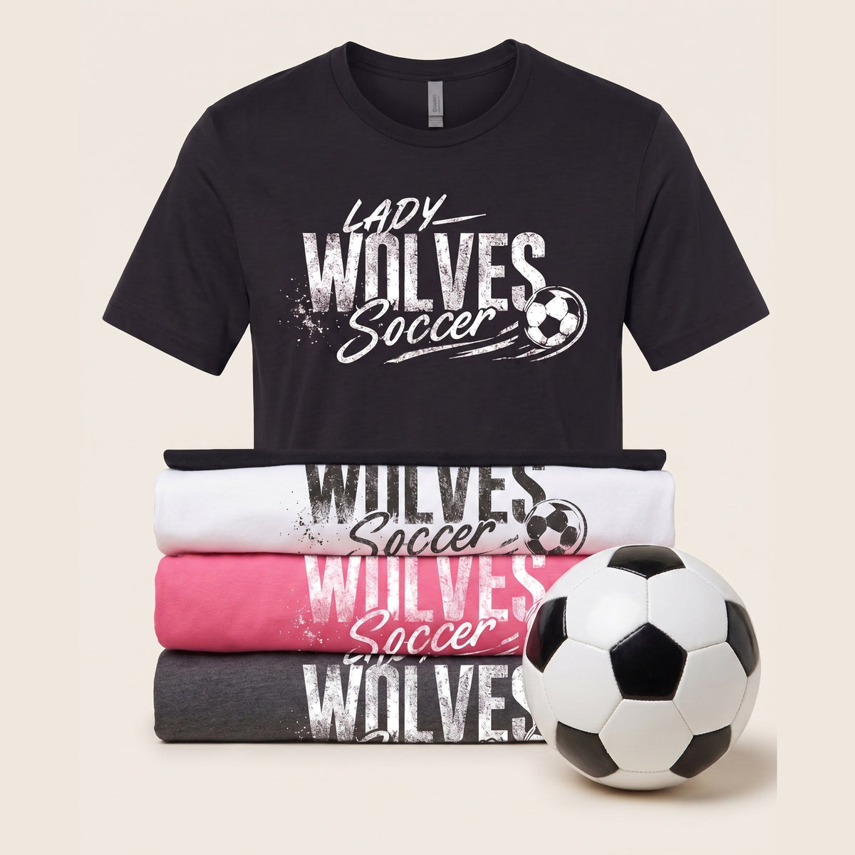 A black short-sleeve graphic T-shirt with white text reading 'Lady Wolves Soccer' and a soccer ball graphic, displayed above three folded T-shirts in white, pink, and dark gray, with a black-and-white soccer ball in front, on a light beige background.