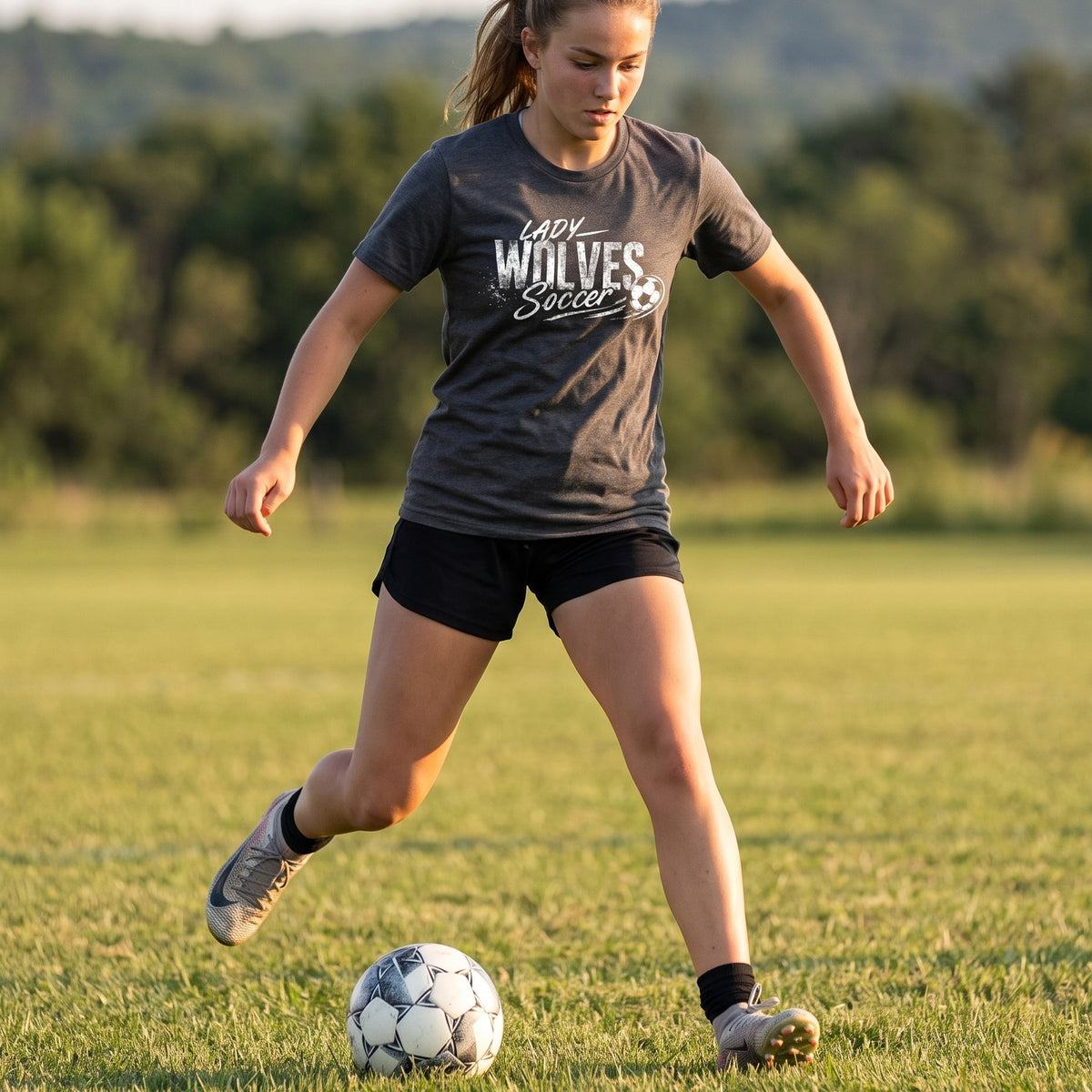 A woman playing soccer on a grassy field wearing a dark gray graphic t-shirt with 'LADY WOLVES Soccer' printed on the front, black athletic shorts, and soccer cleats, with a soccer ball at her feet.